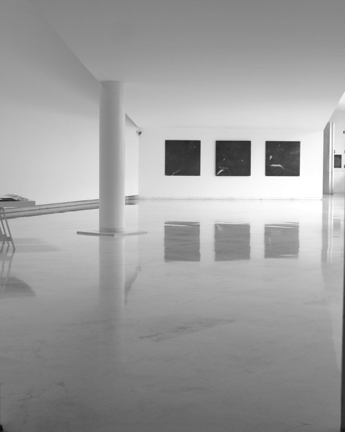 Three dark, square paintings on a white wall in an empty gallery.