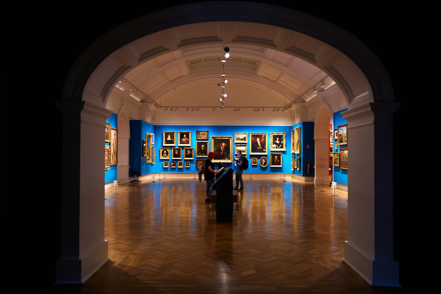 Odwiedzający zwiedzają galerię sztuki z niebieskimi ścianami i obrazami.